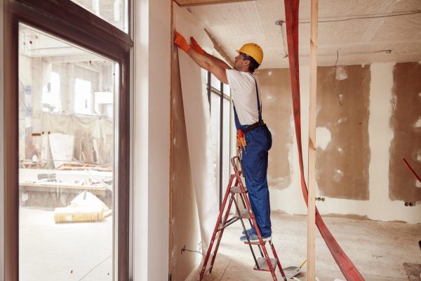 Handsome young man in work overalls standing on ladder and wallpapering wall in house under construction